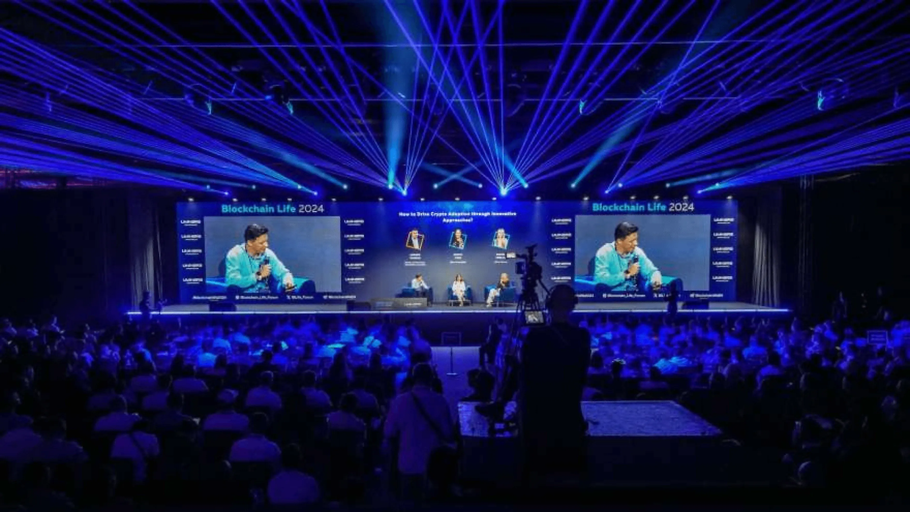 Blockchain Life 2024 conference with industry experts discussing cryptocurrency adoption on a large stage with blue lighting and an engaged audience.
